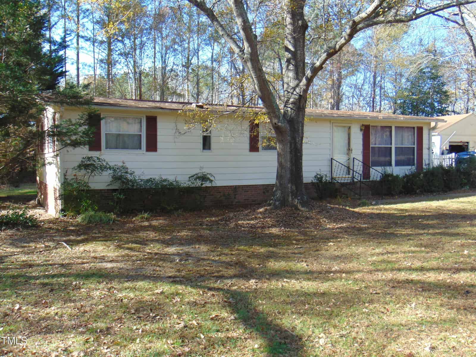 A single-story beige house with a brick foundation, red shutters, and a large front window. A leafless tree stands in the front yard, surrounded by grass and shrubs. The background features more trees and a clear blue sky.