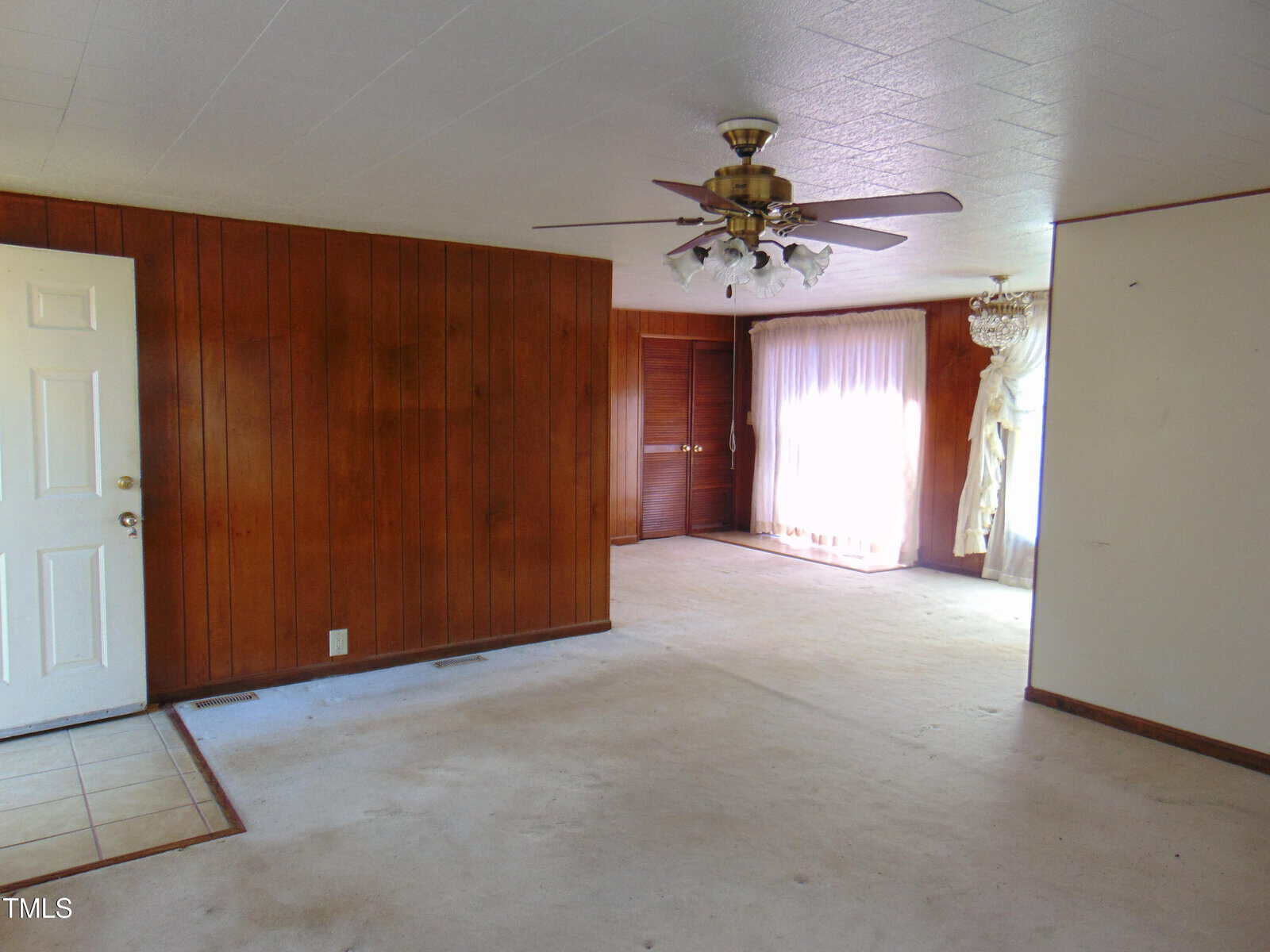 Empty room with wood-paneled walls, white ceiling, and white carpet. There's a ceiling fan with lights, a closed white door on the left, and large windows with white curtains on the right, letting in natural light.