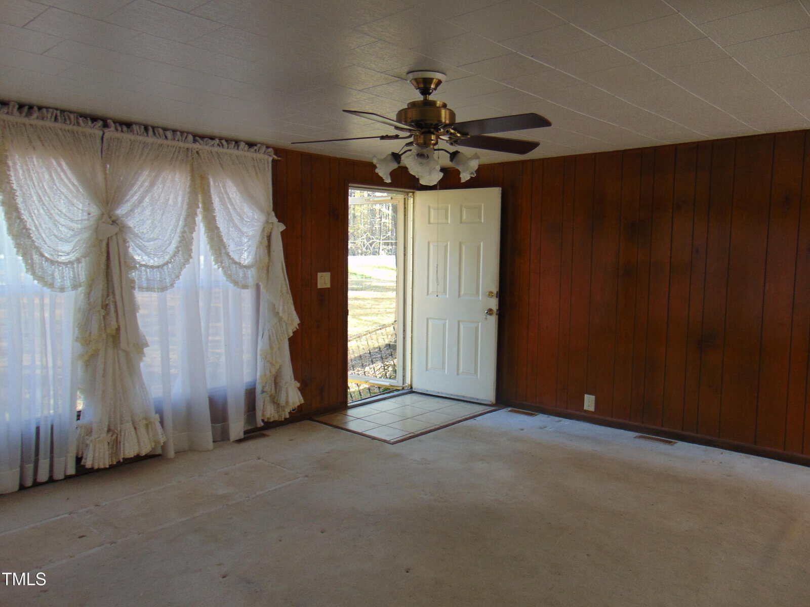 A room with wood-paneled walls, white curtains on a large window, a white door partially open, and a ceiling fan with lights. The floor is bare and there is natural light streaming in through the window.