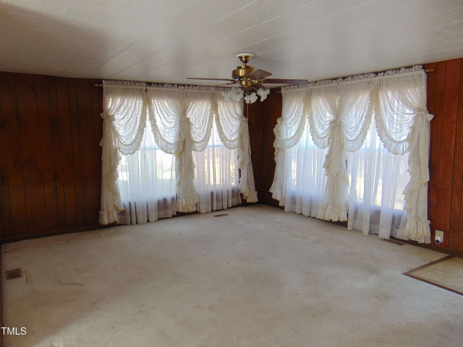 A living room with brown wood-paneled walls, white curtains with lace details, and a ceiling fan. The room is carpeted and filled with natural light from large windows.