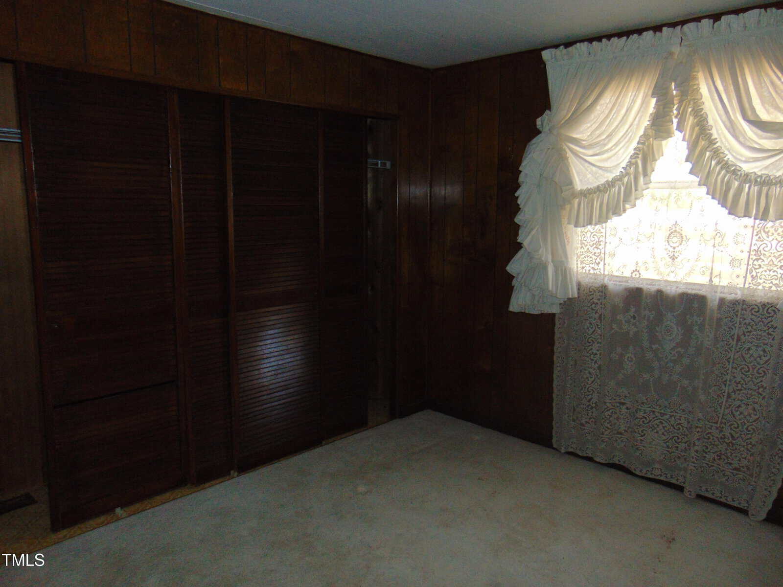 A dimly lit room with dark wood-paneled walls. It features a closed wooden closet on one side and a window with ornate lace curtains and draped valances on the other. The carpet is light-colored.