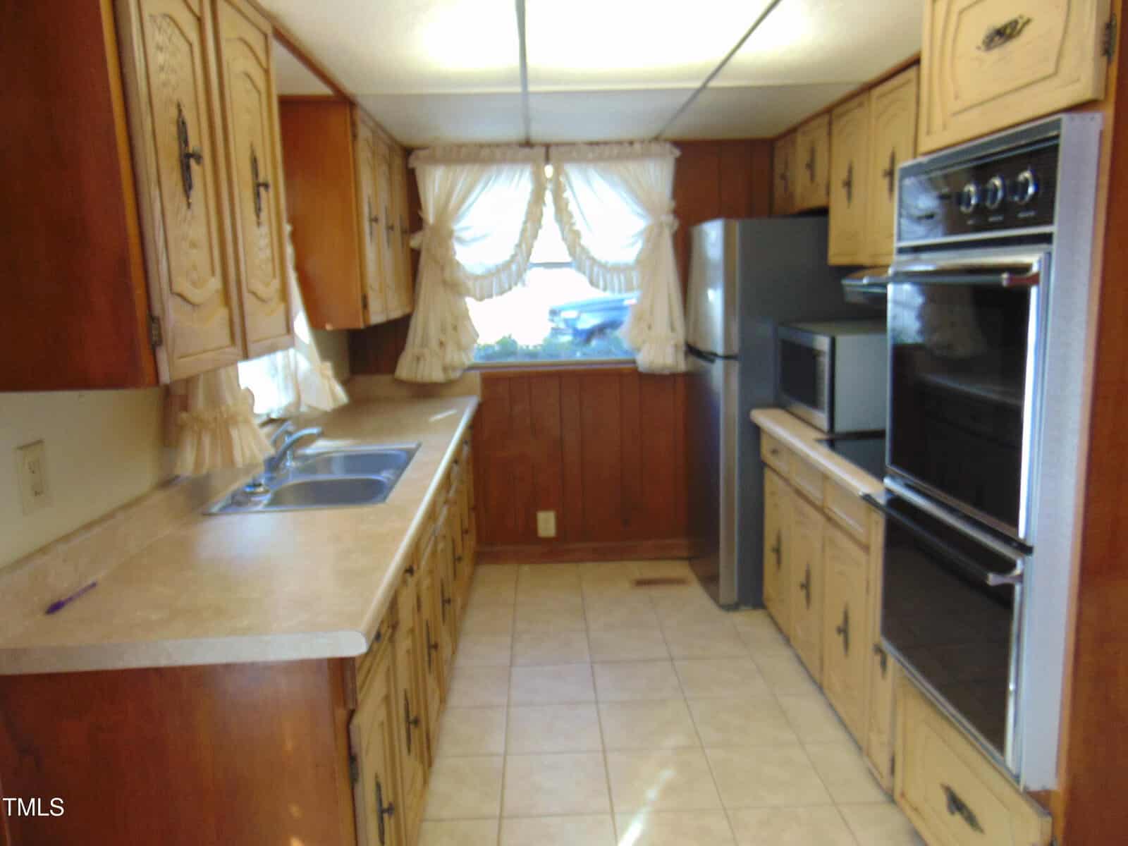 A narrow kitchen with wooden cabinets, a double stainless steel sink, and a beige countertop. A silver refrigerator stands next to a double wall oven. Bright curtains adorn the window above the sink. The floor has light-colored tiles.