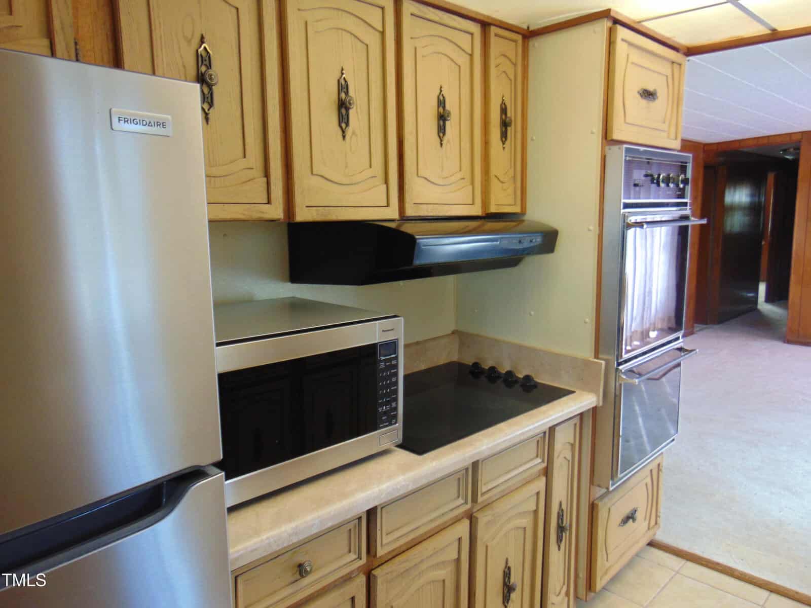 A kitchen featuring wooden cabinets, a stainless steel refrigerator, a modern microwave, a black stovetop with four burners, and a double wall oven with chrome finish. The space is well-lit, highlighting the natural wood tones.