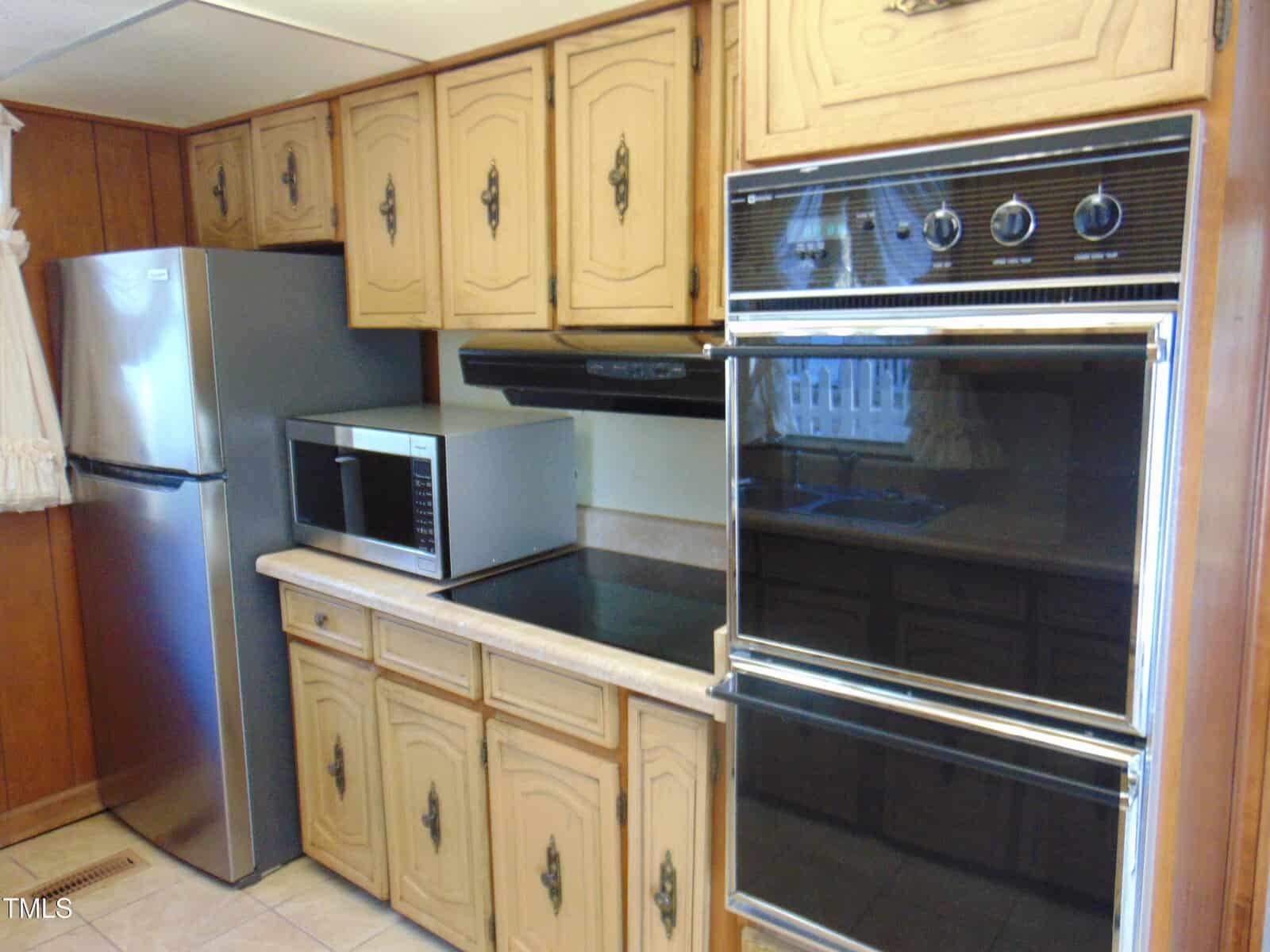A kitchen with wooden cabinets, a stainless steel refrigerator, a microwave on the counter, and a double wall oven. The countertops are beige, and the flooring is tiled. A small window with a curtain is visible.