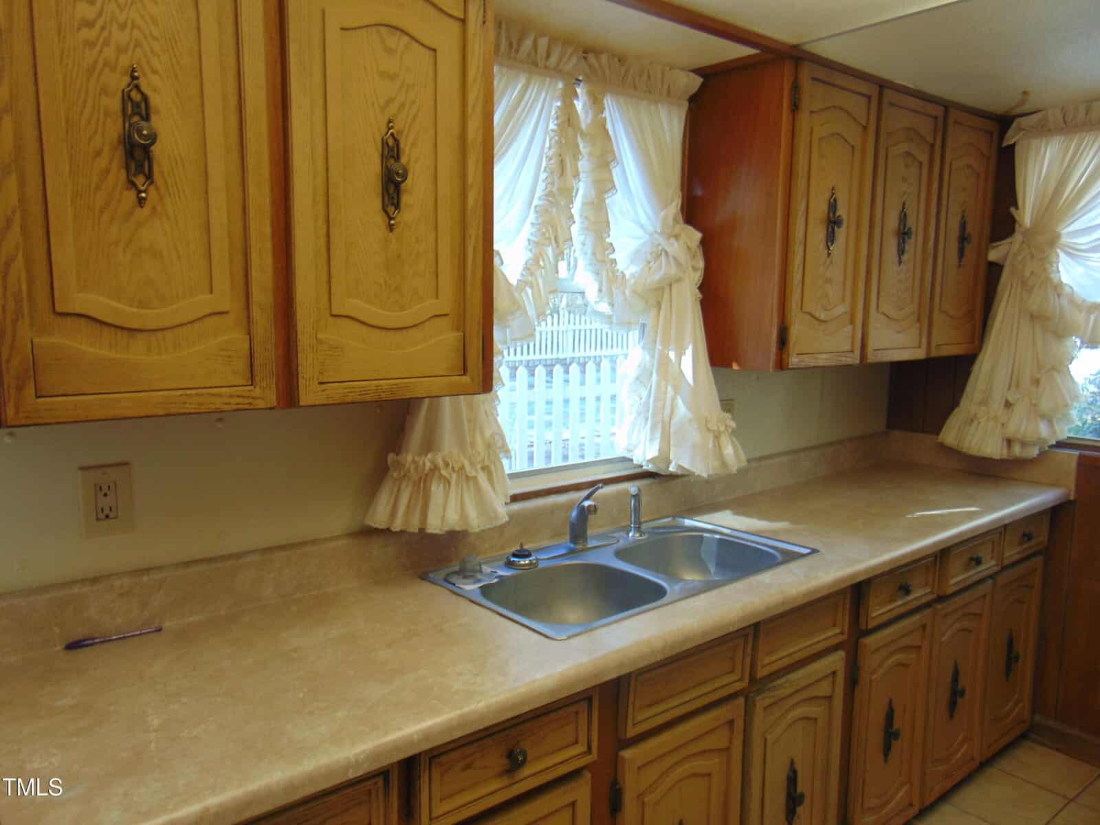A retro kitchen features wooden cabinets, a double stainless steel sink, and beige countertops. Frilly white curtains adorn the windows, allowing natural light to illuminate the space.