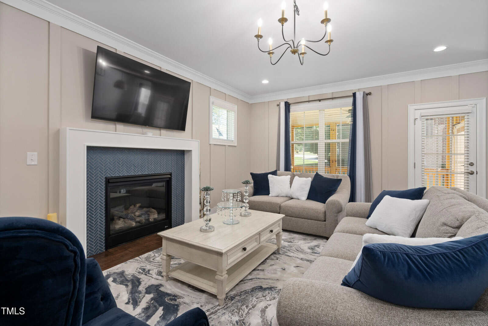 Living room with a modern design, featuring a mounted TV above a fireplace. The space includes a marble-top coffee table, gray and navy seating, and a patterned area rug. Natural light streams in through windows with curtains. A chandelier hangs from the ceiling.