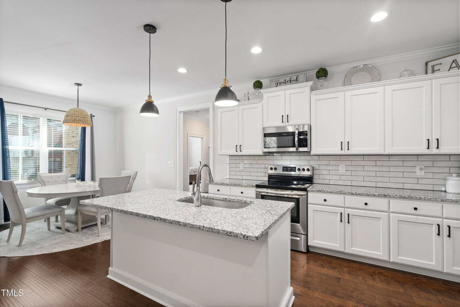 Modern kitchen with a granite island and stainless steel appliances, including a stove and microwave. White cabinets with black handles line the walls. A dining area with a round table and chairs is visible near large windows with blue curtains.