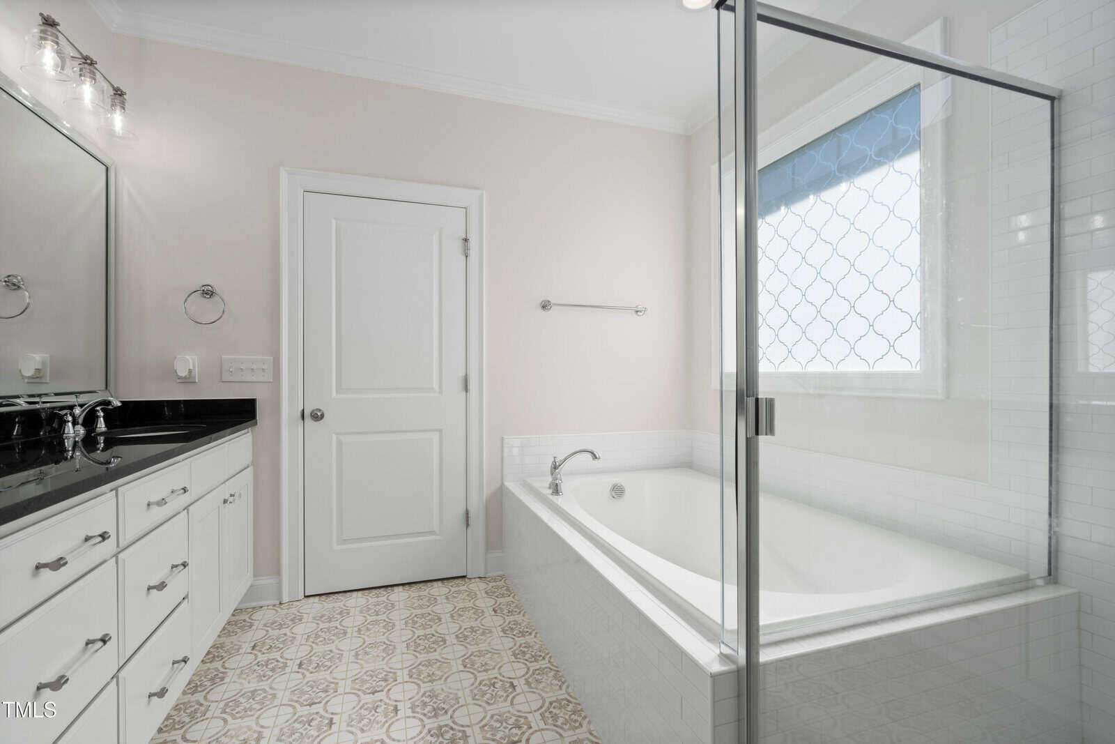 A modern bathroom featuring a large glass-enclosed bathtub with a tiled surround, white cabinets with black countertops, a wall mirror, and patterned floor tiles. Natural light enters through a window with a decorative privacy film.