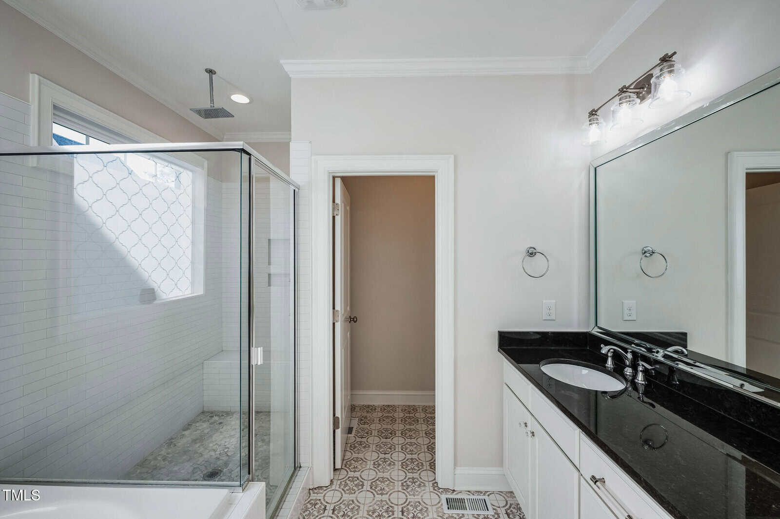 Modern bathroom with a glass-enclosed shower on the left, featuring white subway tiles. To the right, a black granite countertop with dual sinks under a large mirror and two light fixtures. The floor has patterned tiles, and a closed door is in the center.
