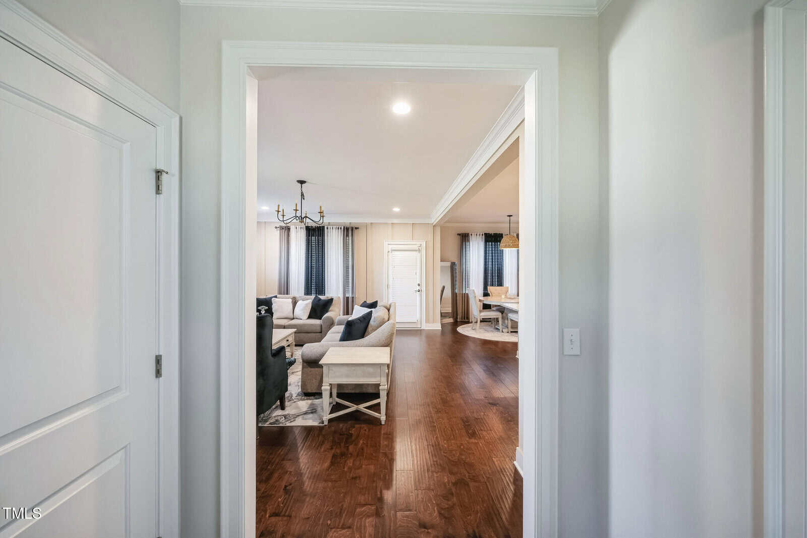 View from a hallway into a spacious living room with wooden floors. The room features a seating area with sofas, a chandelier, and large windows with curtains. Adjacent is a dining area with a table and chairs. Walls are painted in neutral tones.