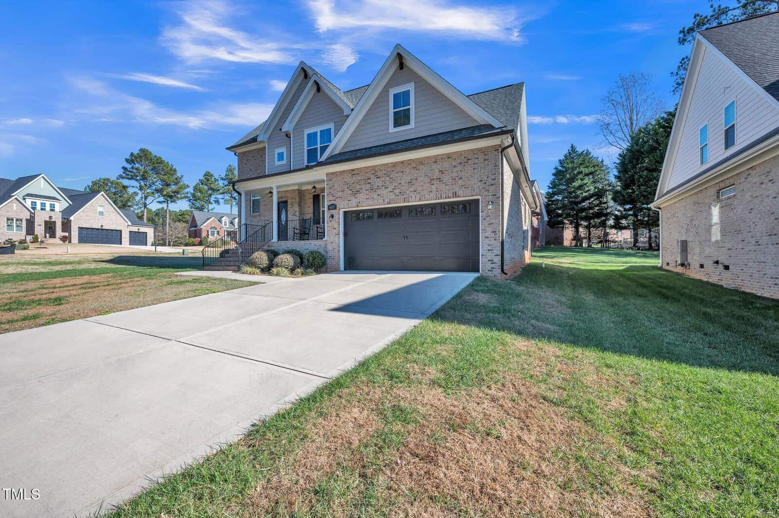A modern two-story brick house with a covered porch and a two-car garage. The driveway is concrete, and the lawn is neatly maintained. Nearby, there are similar houses under a clear blue sky.
