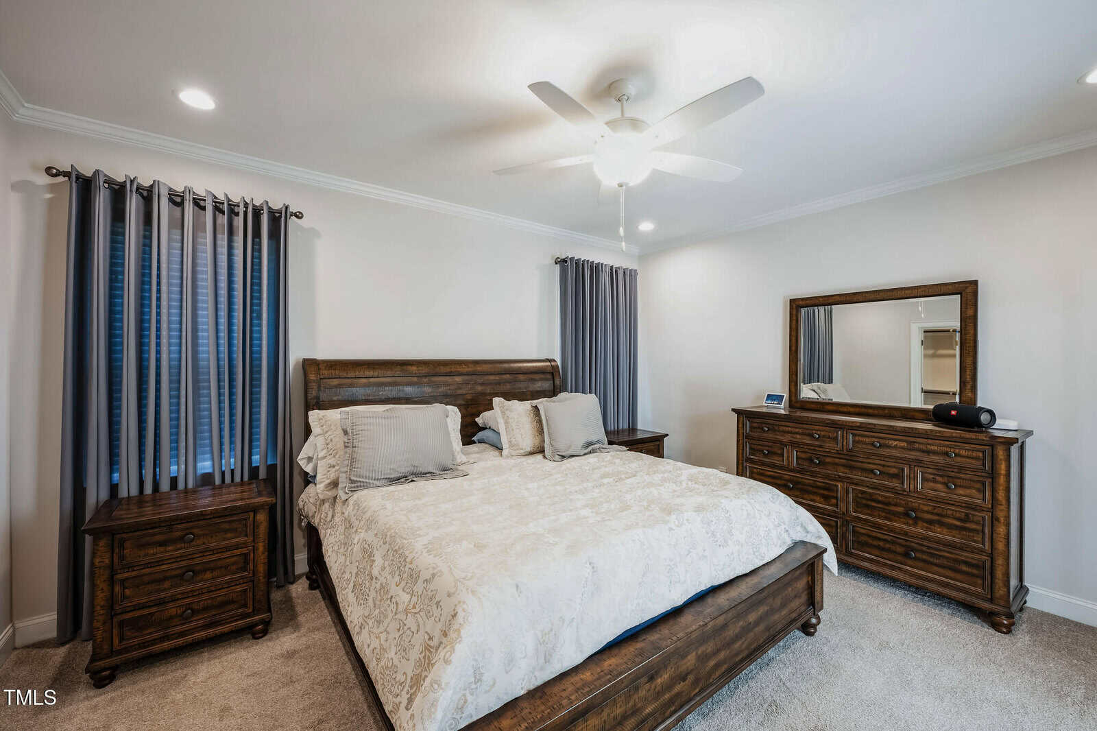 A cozy bedroom with a large wooden bed, ornate headboard, and light-colored bedding. Flanked by two wooden nightstands, it features a dresser with a mirror. Gray curtains cover the windows, and a ceiling fan is above. The carpet is beige.