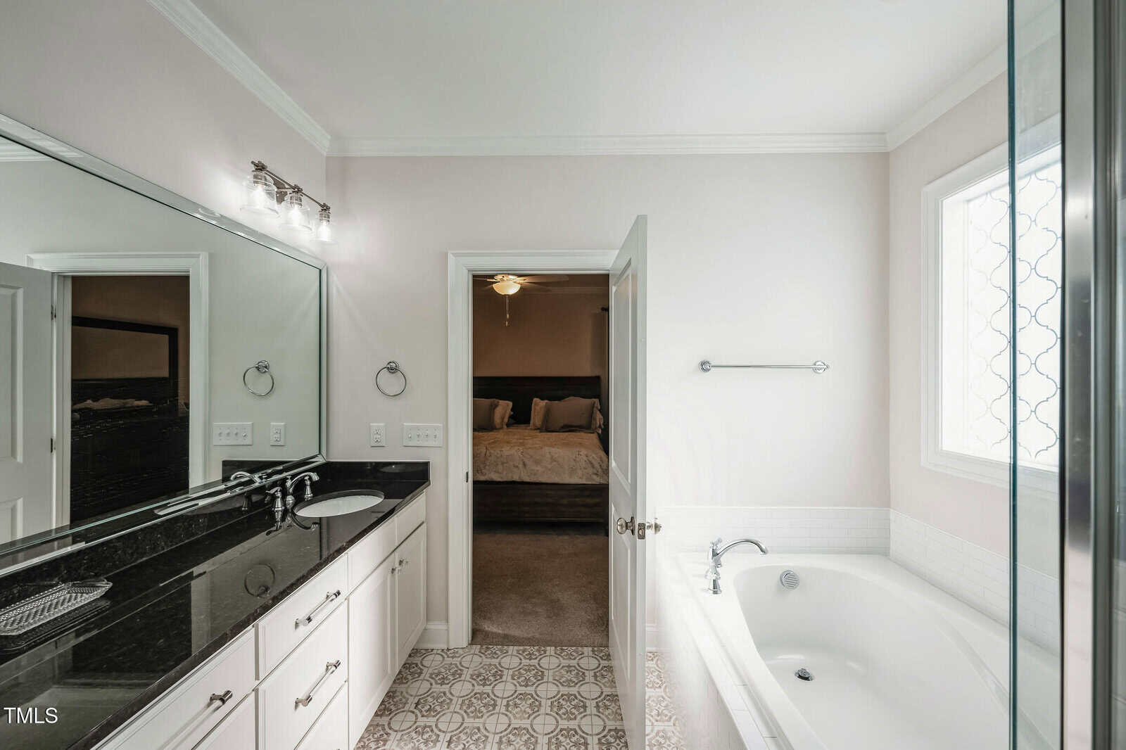 A modern bathroom with a black granite double sink vanity, large mirror, and bright lighting. There's a bathtub with a window above it, and an open door leading to a bedroom with a bed in view. The floor features patterned tiles.