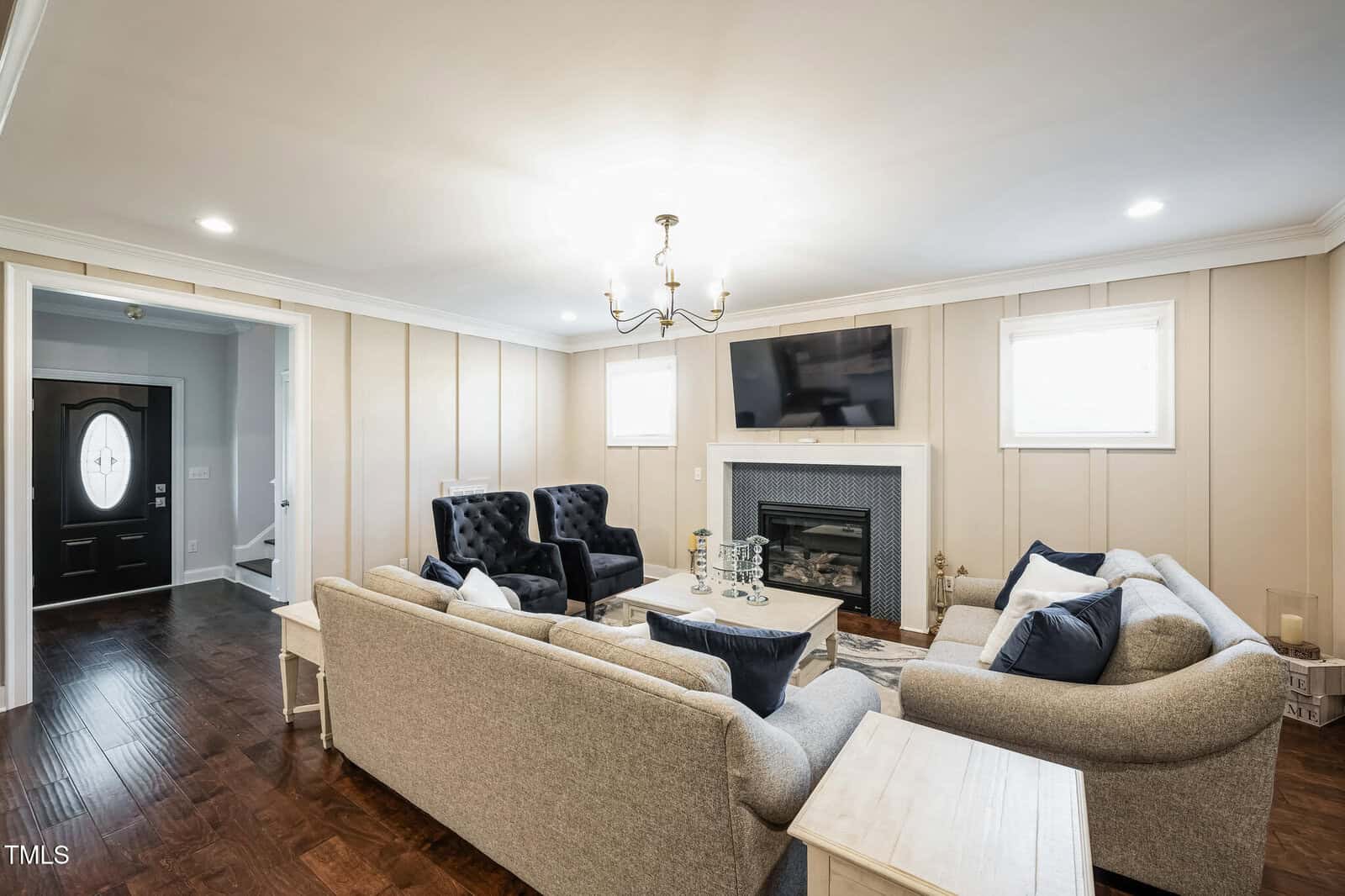 A cozy living room with a modern chandelier, two comfortable gray sofas, black armchairs, and a fireplace. A flat-screen TV is mounted above the fireplace. The room features wooden flooring and a circular coffee table at the center.
