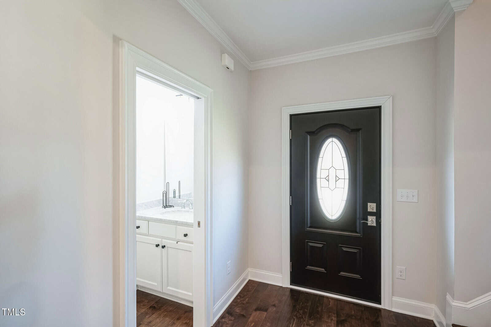 A hallway with a dark front door featuring an oval glass window. To the left is an open doorway leading to a bathroom, visible with a sink and faucets. The flooring is dark wood, and the walls are painted light gray.