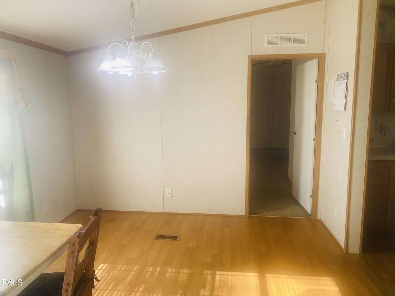 A sunlit room with wooden flooring, featuring a hanging light fixture above a dining area. A wooden chair is partially visible on the left. The walls are light-colored with a doorway leading to a darker room on the right.