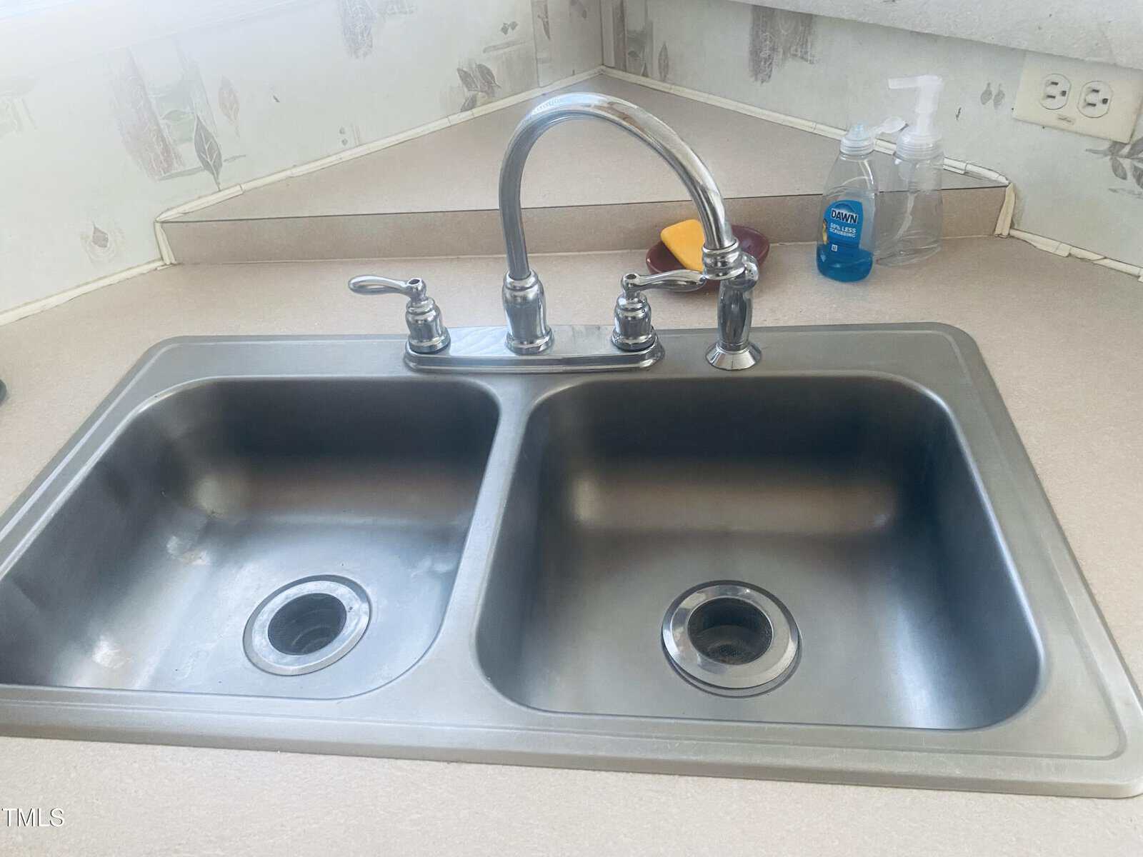 A stainless steel double kitchen sink with a chrome faucet. There are cleaning supplies, including a hand soap dispenser and a sponge, on the countertop. The backsplash is decorated with light-colored wallpaper.