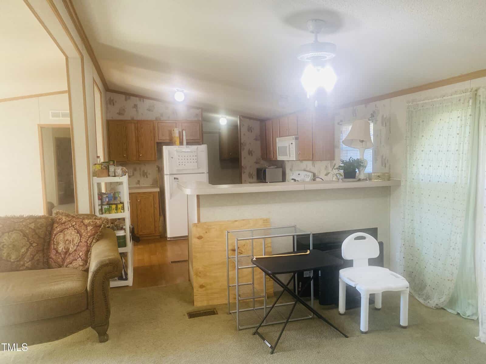 A cozy living room with beige carpeting, featuring a brown sofa on the left. An open kitchen with wooden cabinets and white appliances is in the background. A small table and a child's chair are positioned near a curtained window on the right.