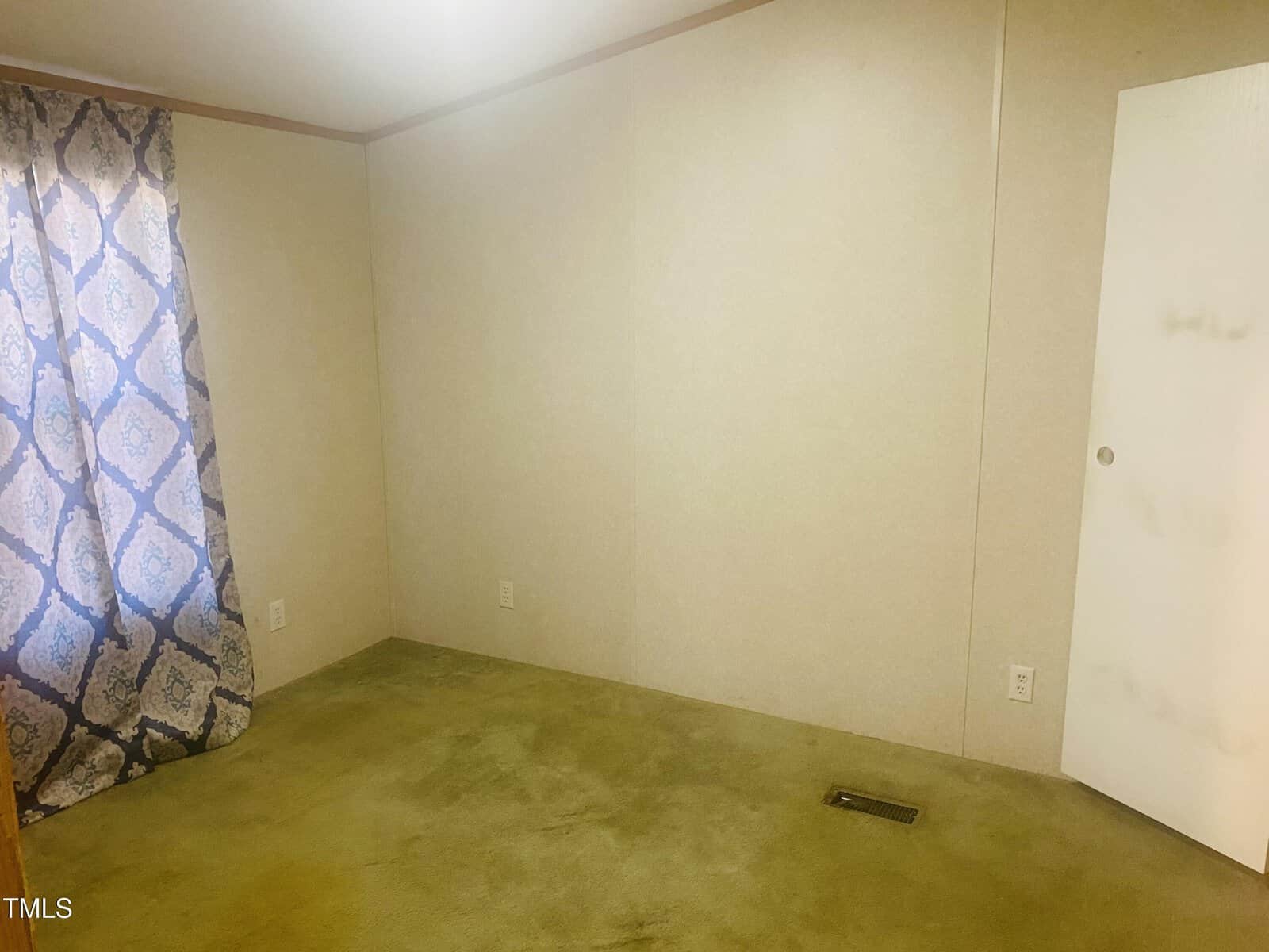 An empty room with beige walls and a closed white door. A single window is covered by a curtain with a blue and white pattern. The floor is carpeted in a light green color. The room appears bare and undecorated.