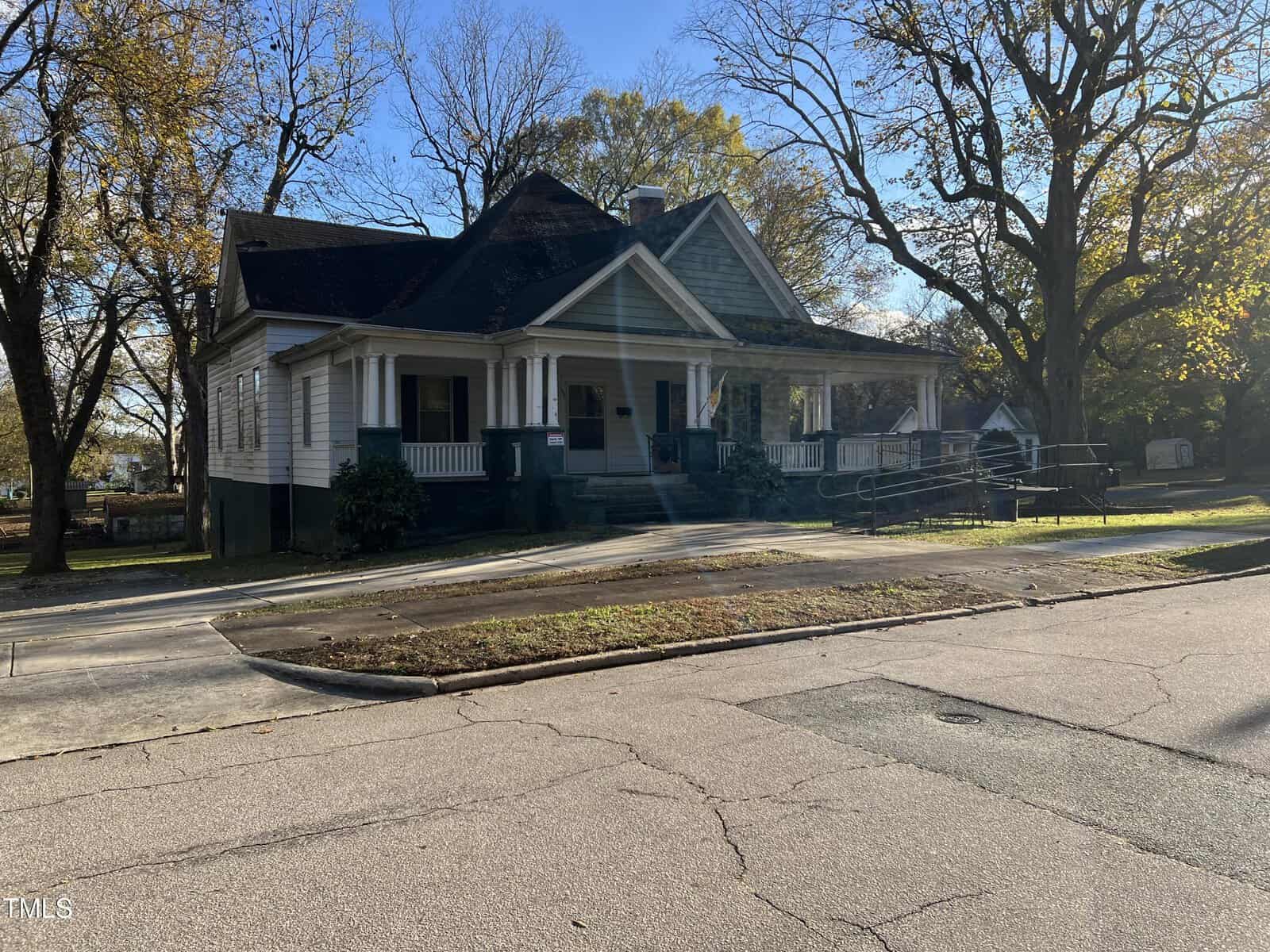 A white, single-story house with a gabled roof and a wrap-around porch stands in a leafy neighborhood. The yard is mostly grass, and there is a paved driveway. Leafless trees surround the house under a clear blue sky.