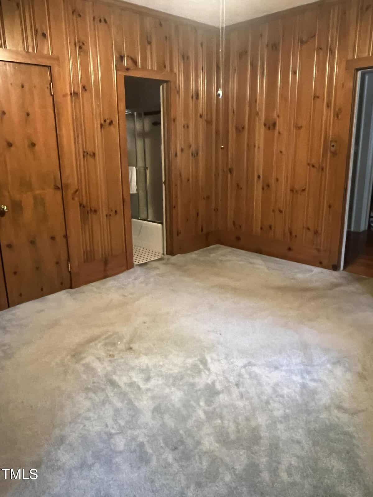 Room with wood-paneled walls and light-colored carpet. There is an open door leading to a bathroom with a visible shower. Two doorways are present, one to the left and one to the right. The room is empty and well-lit.