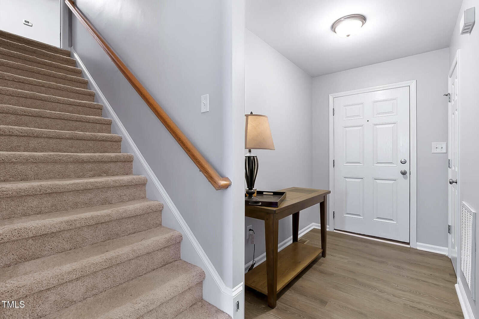 A hallway with a front door, a wooden table with a lamp and books, and carpeted stairs with a wooden handrail leading upward. Walls are light gray; the floor is wood in the hallway and carpeted on the stairs.