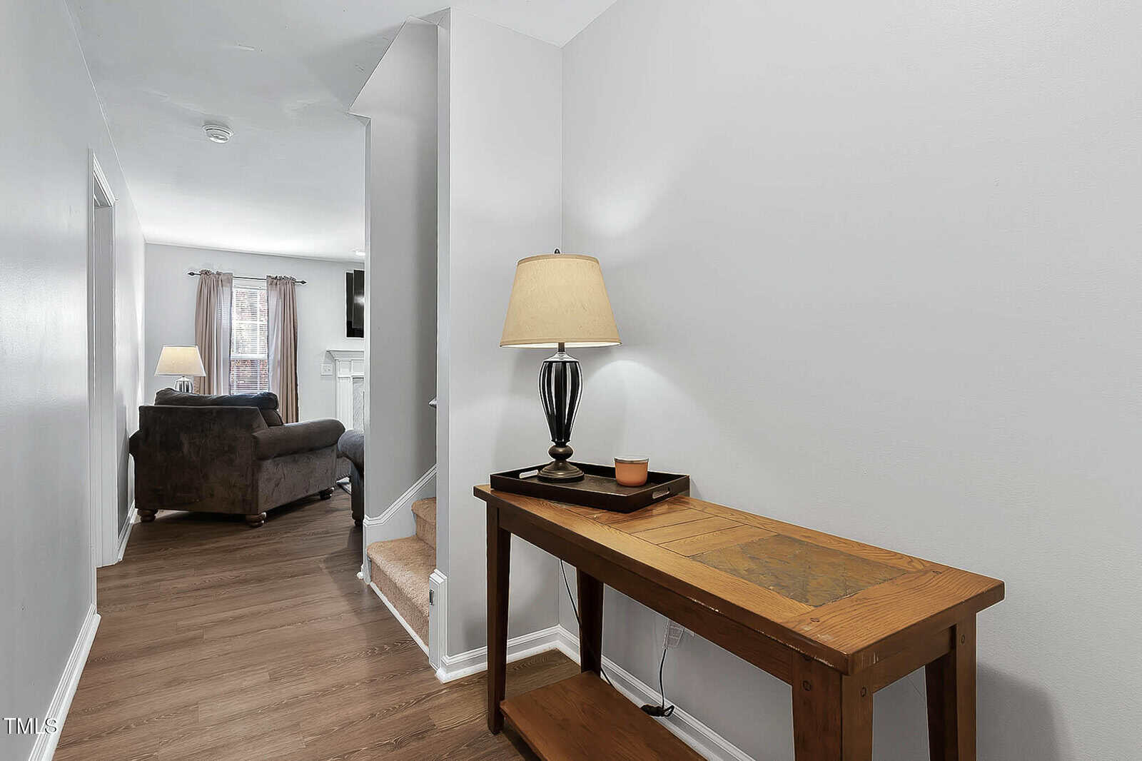 A wooden console table with a lamp and a small dish stands in a hallway with wood floors. A living room with a couch and window curtains is visible in the background.