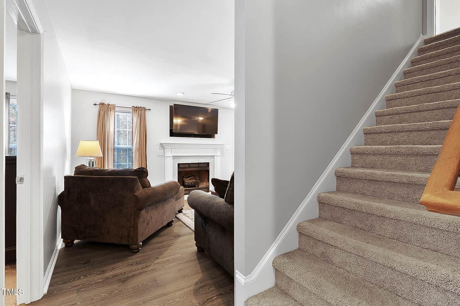A cozy living room with brown sofas, a fireplace, TV, and lamp is visible on the left. On the right, carpeted stairs lead upward, with neutral-colored walls throughout the space.