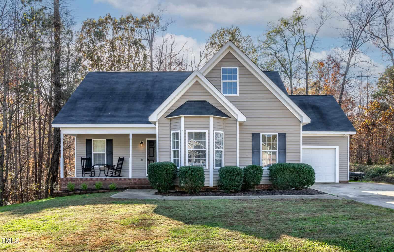 A beige single-family house with white trim, a covered front porch with black rocking chairs, attached garage, front-facing gable, bay window, manicured bushes, and a green lawn bordered by trees.
