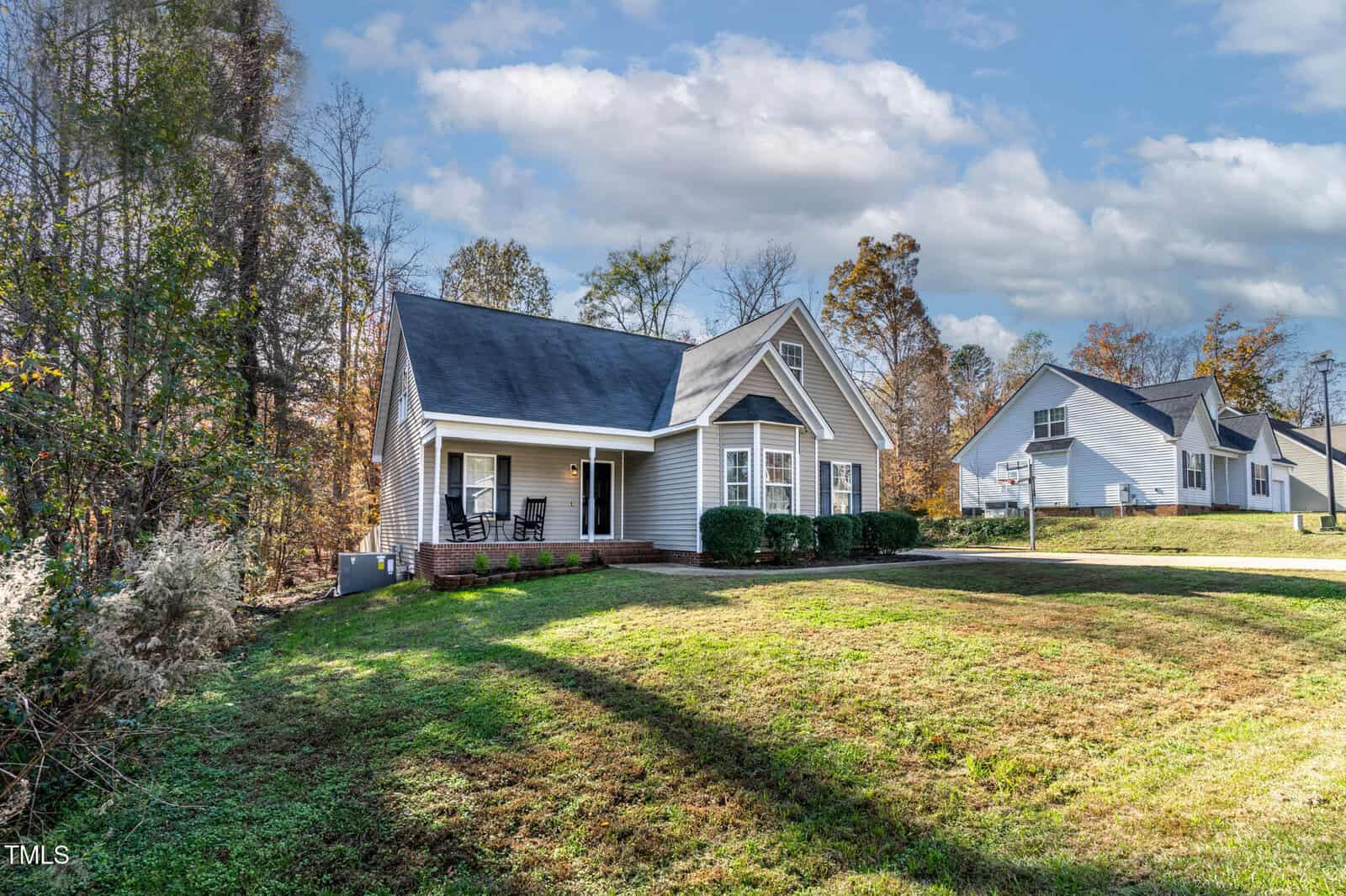 A single-story beige house with black roof shingles and a covered front porch sits on a grassy lawn. Trees border the property on the left, and another house is visible in the background. The sky is partly cloudy.