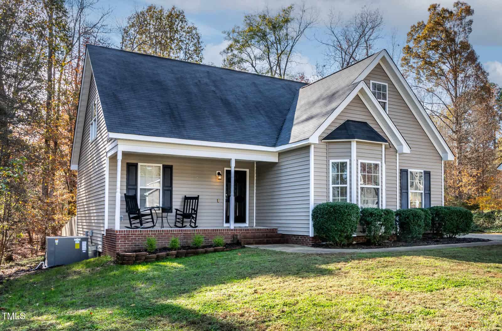 A single-story house with gray siding, white trim, a dark roof, and a front porch with two black rocking chairs. There are bushes along the front, and trees with fall foliage in the background.