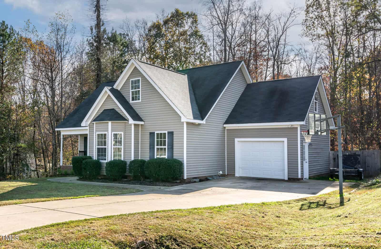 A gray, single-story house with white trim, a one-car garage, and a basketball hoop in the driveway. The yard is grassy with trimmed bushes along the front and trees in the background.