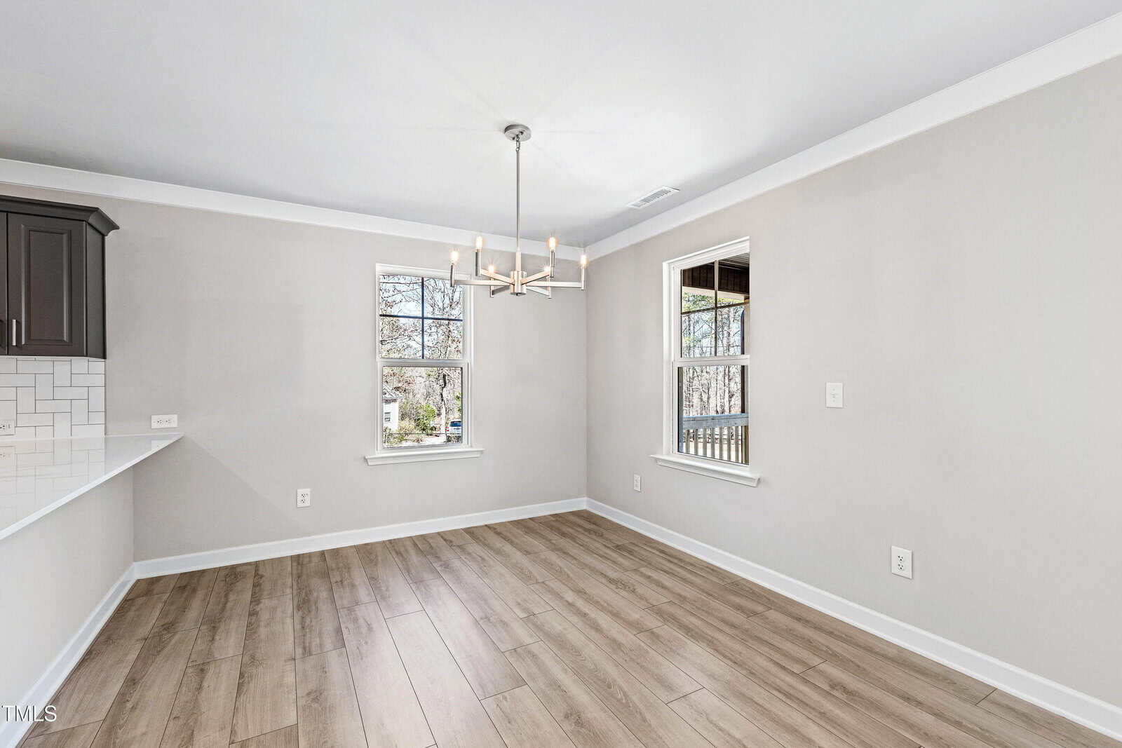 A bright, empty dining area with light wood flooring, two windows, gray walls, and a modern chandelier. Part of a kitchen counter and white subway tile backsplash are visible on the left.