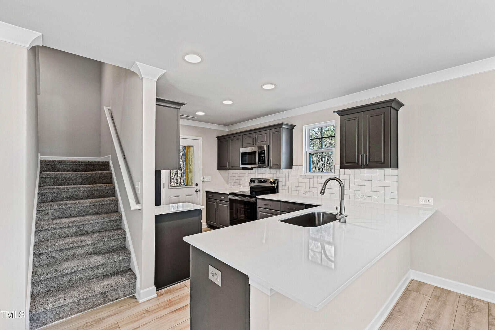 Modern kitchen with white countertops, dark cabinets, white subway tile backsplash, built-in sink, and stainless steel appliances. Carpeted stairs are visible on the left side, and light wood flooring runs throughout.