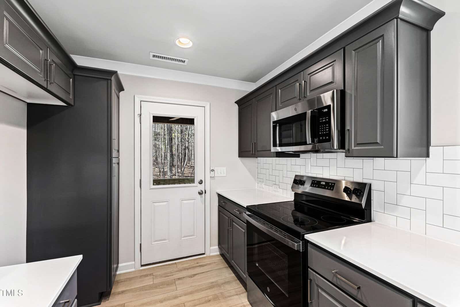 A modern kitchen with dark gray cabinets, stainless steel appliances, a white subway tile backsplash, light wood flooring, and a white door with a window leading outside.