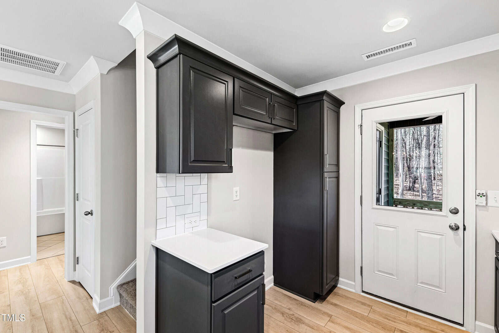 A modern kitchen corner with dark cabinets, white tile backsplash, light wood flooring, and a white exterior door with a window. A small countertop is visible beneath the cabinets.