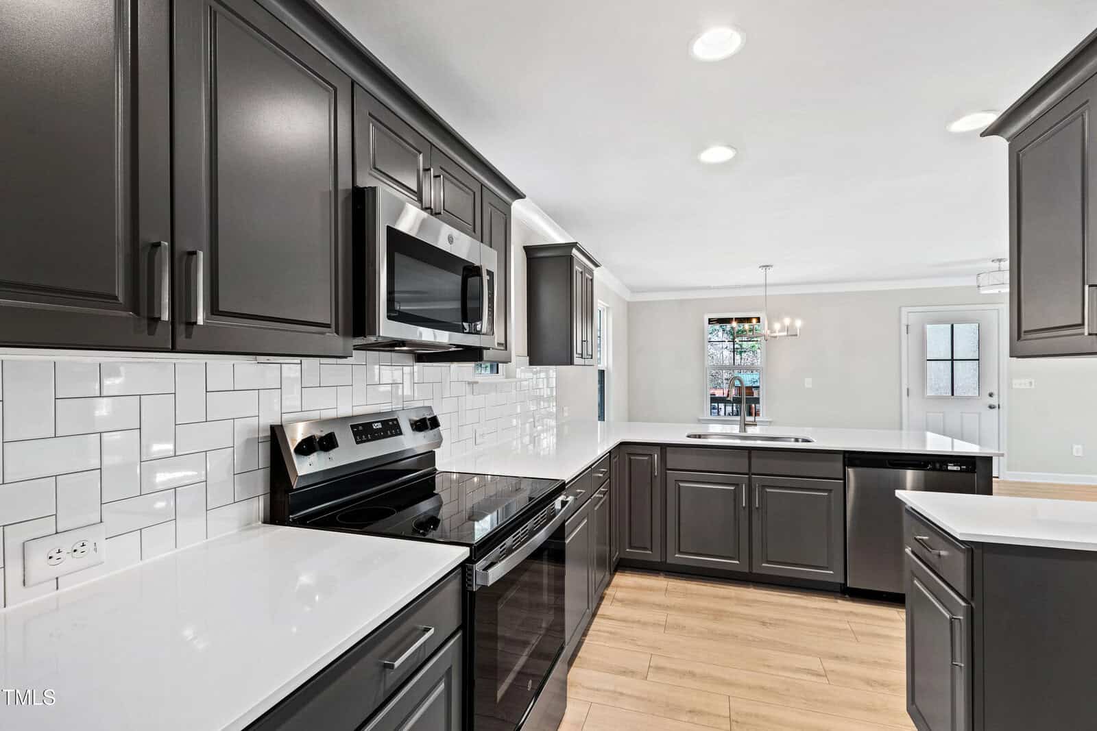 A modern kitchen with dark gray cabinets, white quartz countertops, a white subway tile backsplash, stainless steel appliances, wood flooring, and recessed ceiling lights. A dining area is visible in the background.