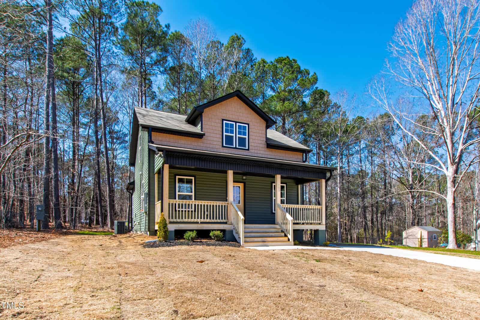 A two-story house with green siding, a covered front porch, and beige trim sits on a yard with sparse grass, surrounded by tall trees under a clear blue sky. A driveway runs along the right side.