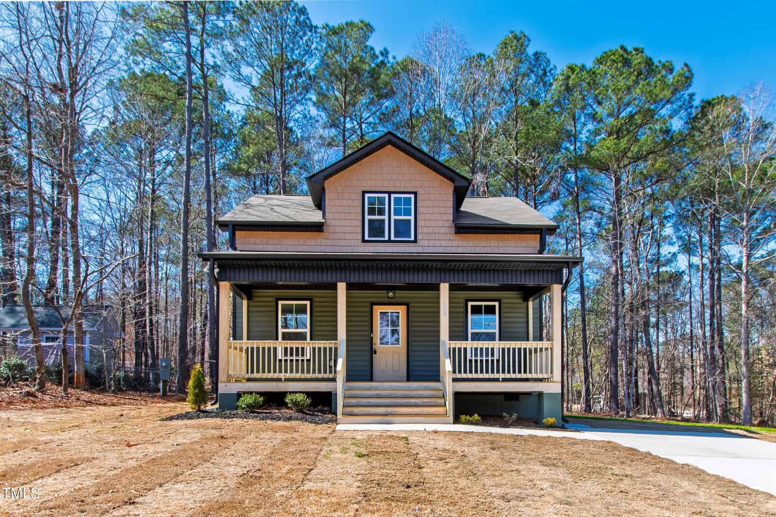 A two-story house with tan siding, a covered front porch, and two dormer windows, set among tall pine trees with a dry lawn and a concrete driveway on a clear day.