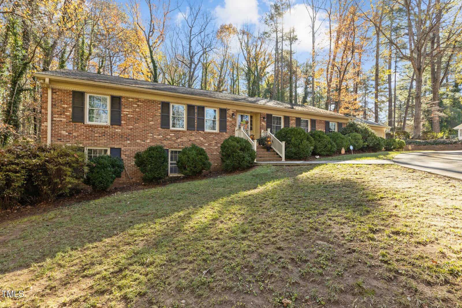 Nestled in a wooded area with autumn trees, the single-story brick house at 161 Pine Cone Drive features multiple windows and a welcoming porch. Its lawn, partially covered with grass, complements the curved driveway on this clear blue sky day in Oxford.