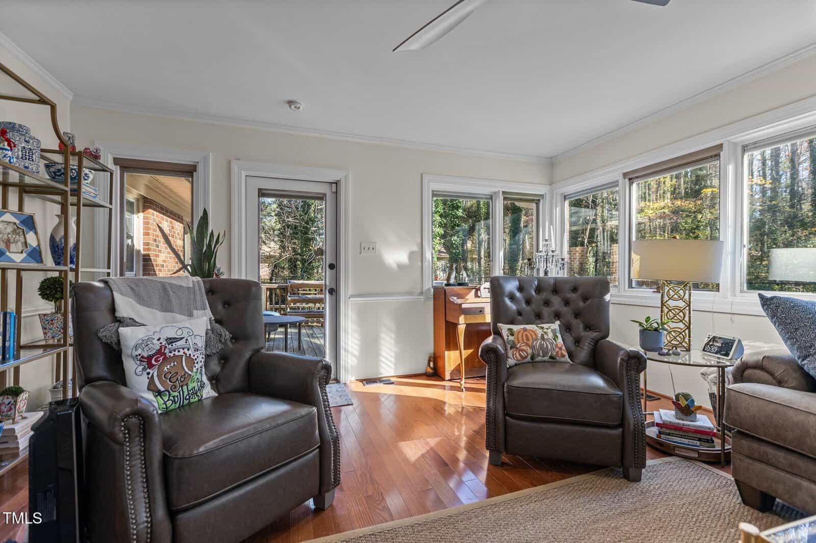 A sunlit living room features two brown leather armchairs and a bookshelf on the left. Large windows offer a view of trees outside. A small desk with a chair is placed near the window. A floor lamp and decor items add to the cozy atmosphere.