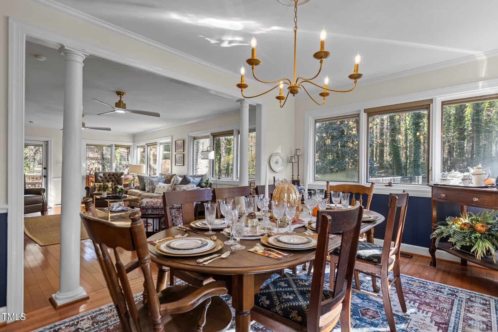 Dining room with a set table featuring elegant dinnerware and glassware. A chandelier hangs above. The room opens into a living area with sofas and a large window view of trees outside. A wooden sideboard is decorated with a floral arrangement.