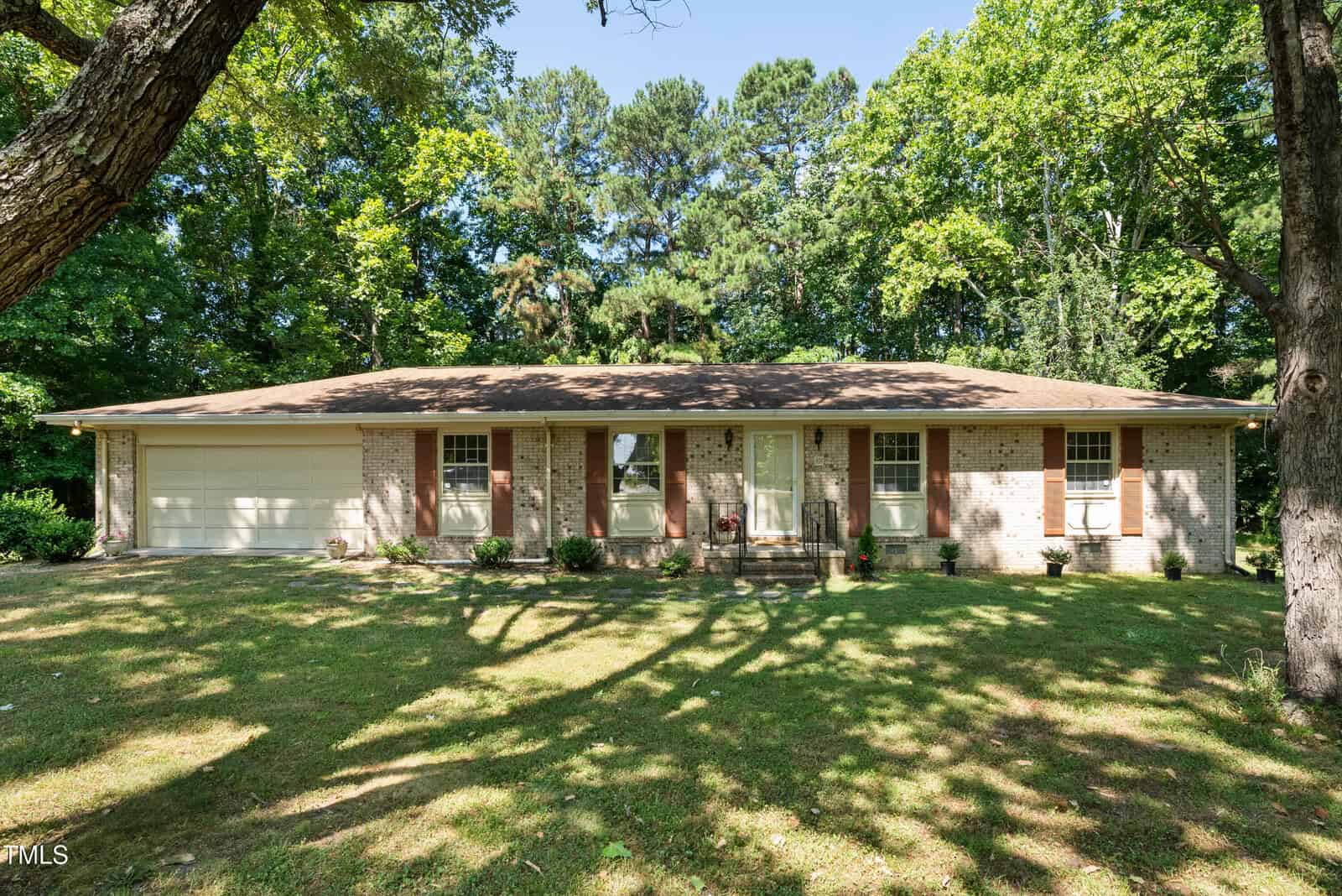 Nestled at 101 Jeffress Drive in Henderson, this single-story brick house features a light brown roof and is surrounded by tall trees. The large garage on the left complements several front windows, while a well-maintained lawn and a few plants adorn the welcoming entrance.