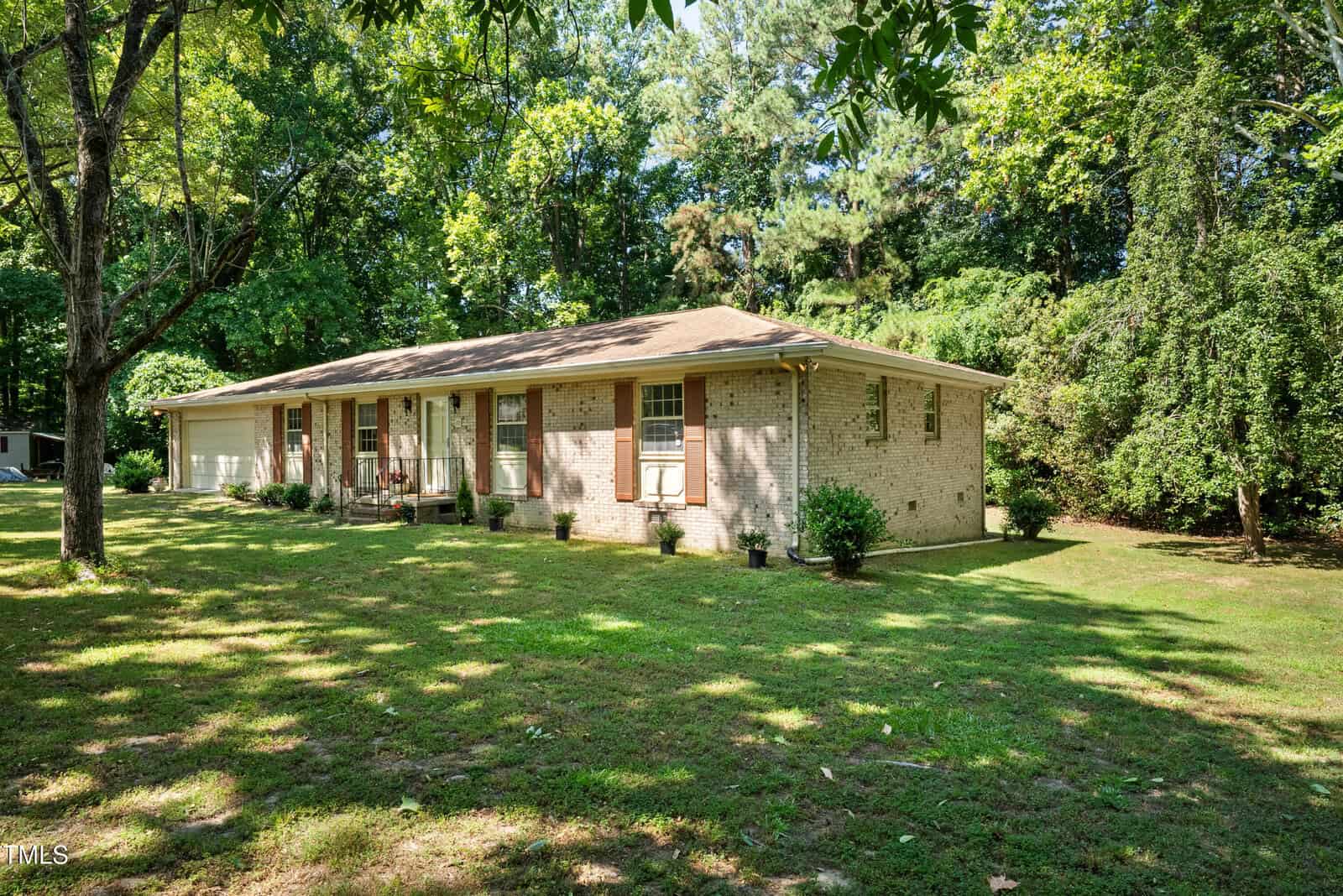 A single-story, light brick house with a brown roof sits amid a lush, green lawn and tall trees. The house features several windows and a front porch with chairs. A clear sky and sunlight enhance the tranquil outdoor setting.
