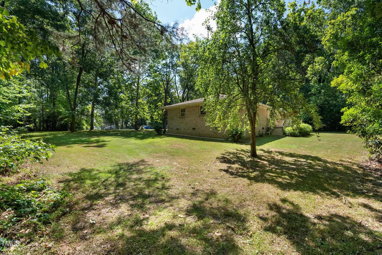 A small house with light-colored siding stands in a wooded area, surrounded by trees. The grassy lawn is sunlit, creating a peaceful and secluded atmosphere. Shadows of trees are cast on the lawn, illustrating a bright, sunny day.