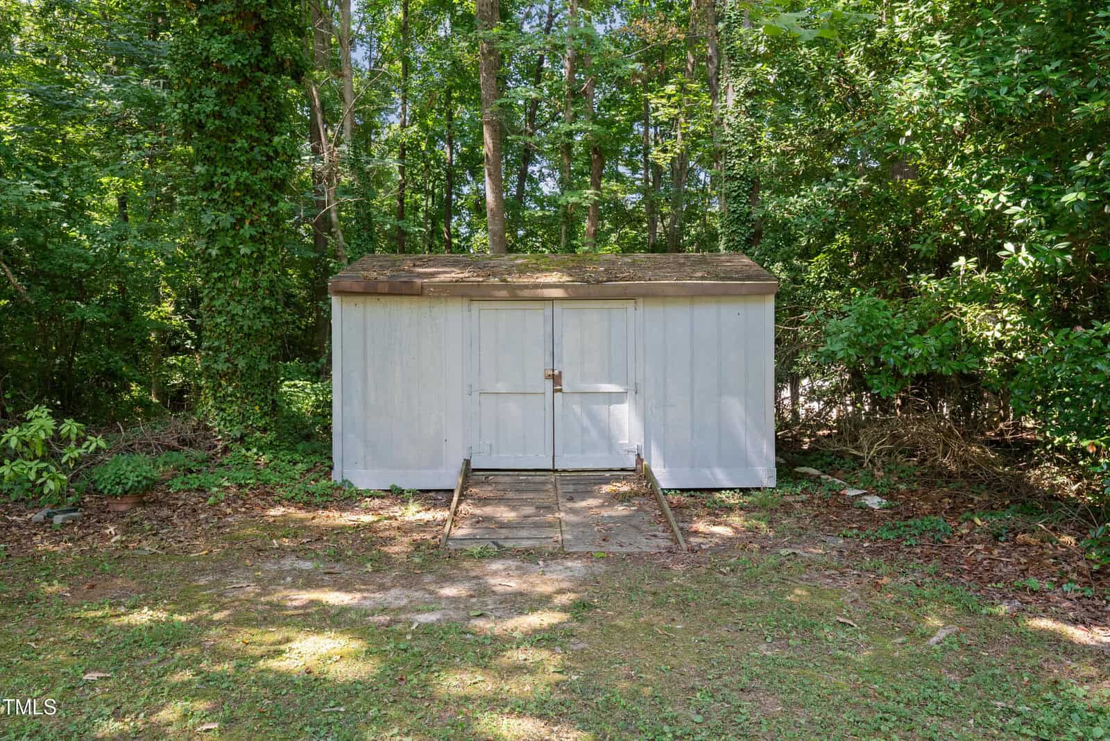 A small, light gray shed with a sloped roof stands in a wooded area. Surrounded by dense trees and foliage, the shed has a front door secured with a metal latch. Sunlight filters through the trees, casting dappled shadows on the ground.