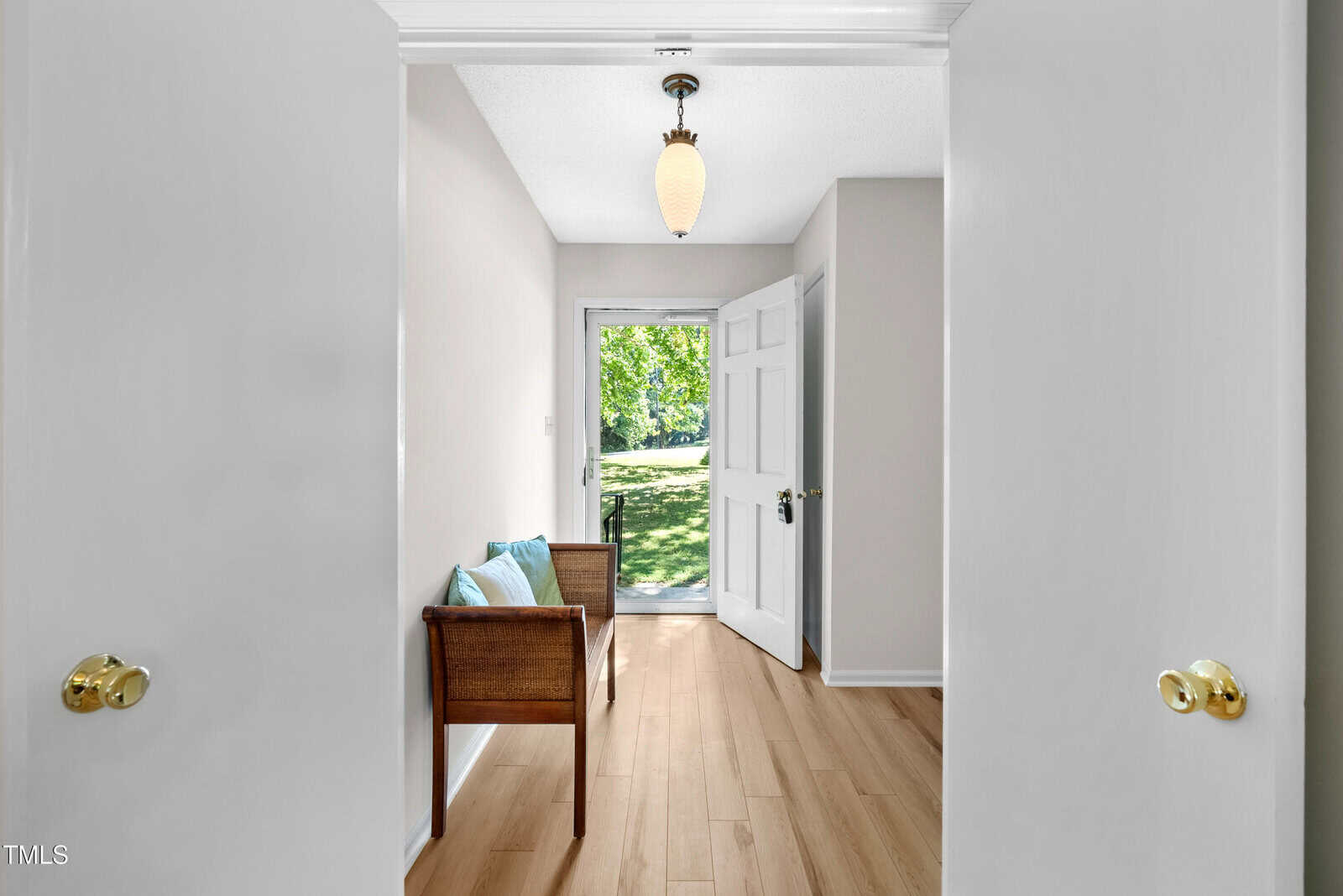 A bright hallway with light wood flooring and white walls leading to a doorway with a view of a green lawn outside. A wicker bench with blue cushions is on the left side, and a hanging light fixture is above.