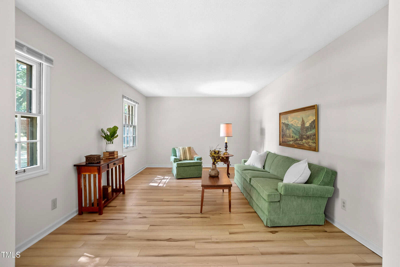 A minimalist living room with wooden floors, a green sofa, an armchair, and a wooden coffee table. A table lamp and framed artwork decorate the space. Large windows allow natural light, and a small wooden console table is against the wall.