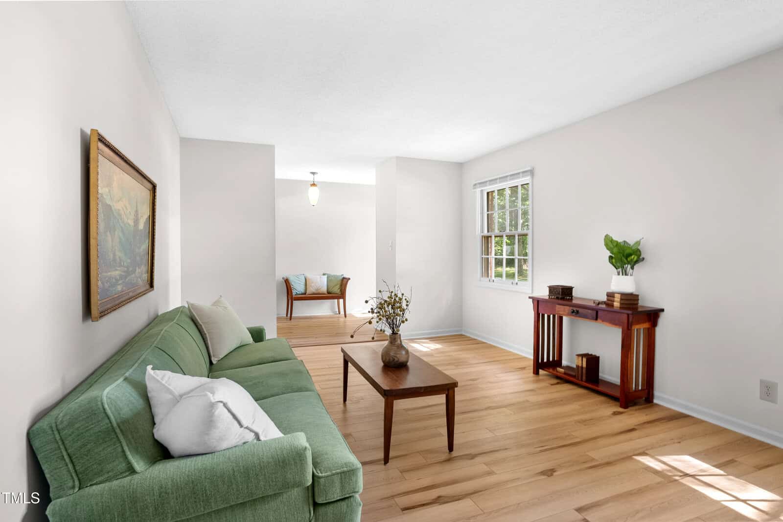 A bright living room with a green sofa, white and gray pillows, and a wooden coffee table with flowers. There's a painting on the wall, and a wooden console with plants by the window. A bench is visible in the adjoining room. Light wood flooring.