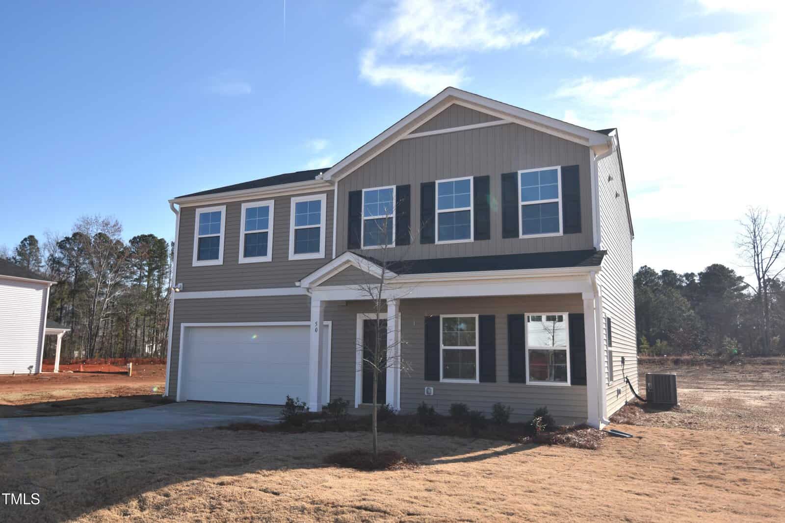 A two-story suburban house with beige siding, white trim, black shutters, and a covered entry porch. The home has a two-car garage and sits on a newly landscaped lawn with a small tree in the front yard.
